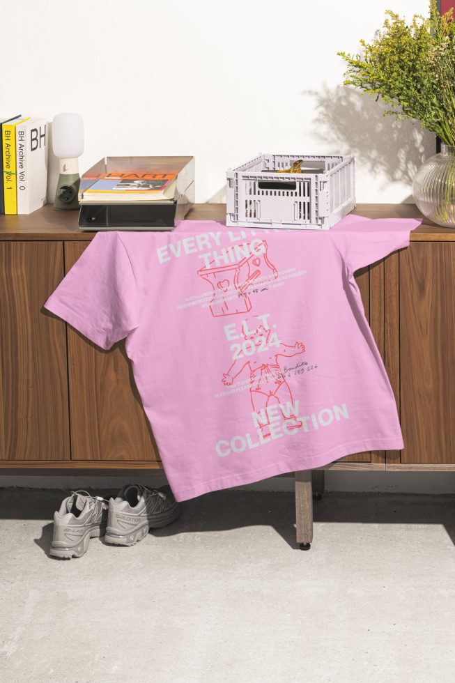 Pink t-shirt with "Every Little Thing" and graphics, draped over a mid-century modern cabinet, next to sneakers. Mockup, graphic design, apparel template.