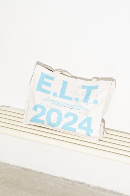 Cream tote bag with blue text E.L.T 2024 on a bench, minimalist design. Ideal for mockups, product displays, and graphic presentations, perfect for designers.