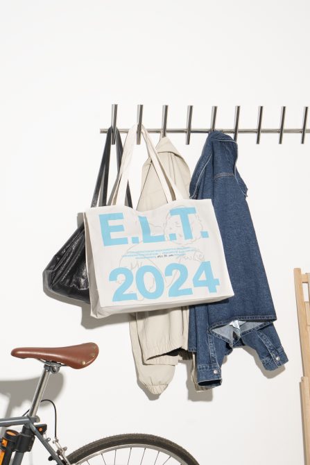 Canvas tote bag mockup with E.L.T 2024 text hanging on a rack alongside a jacket and bike in a minimalist indoor setting design photography.