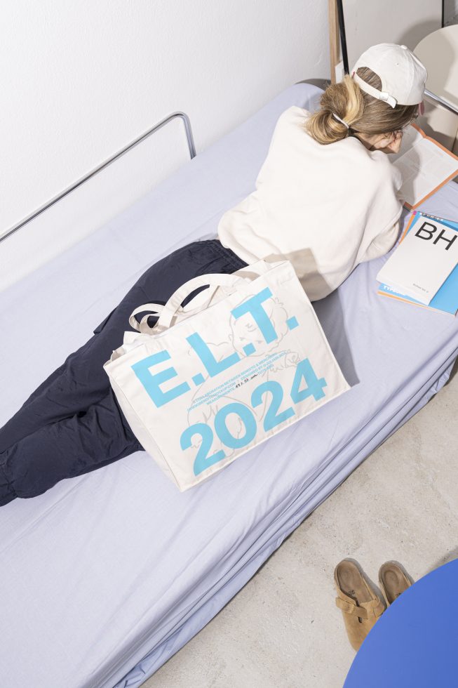 Person reading on bed with tote bag E.L.T 2024 on side, white cap, and casual outfit. Suitable for Mockups, Graphic design, Fashion, Templates, Branding.