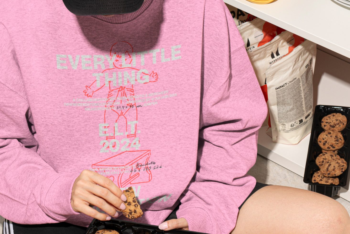 Close-up of a designer wearing a pink graphic sweatshirt while holding a chocolate chip cookie. Ideal for mockups, fashion templates, creative graphics.