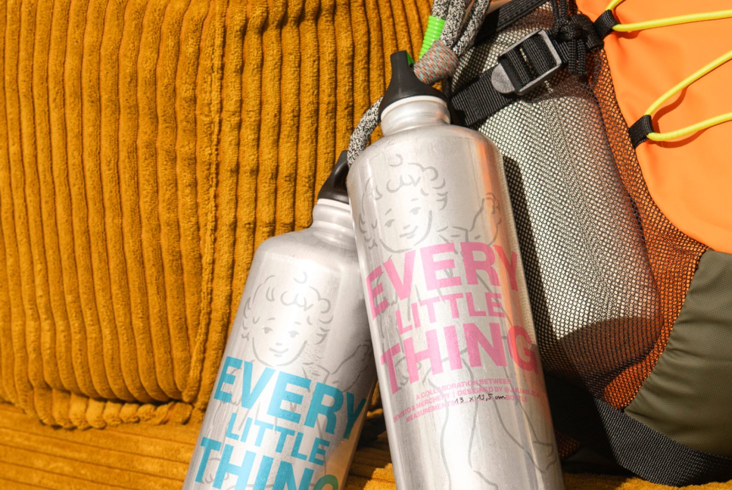 Metal water bottles with pink and blue text reading Every Little Thing leaning on an orange corduroy cushion next to a backpack. Ideal for product mockup.