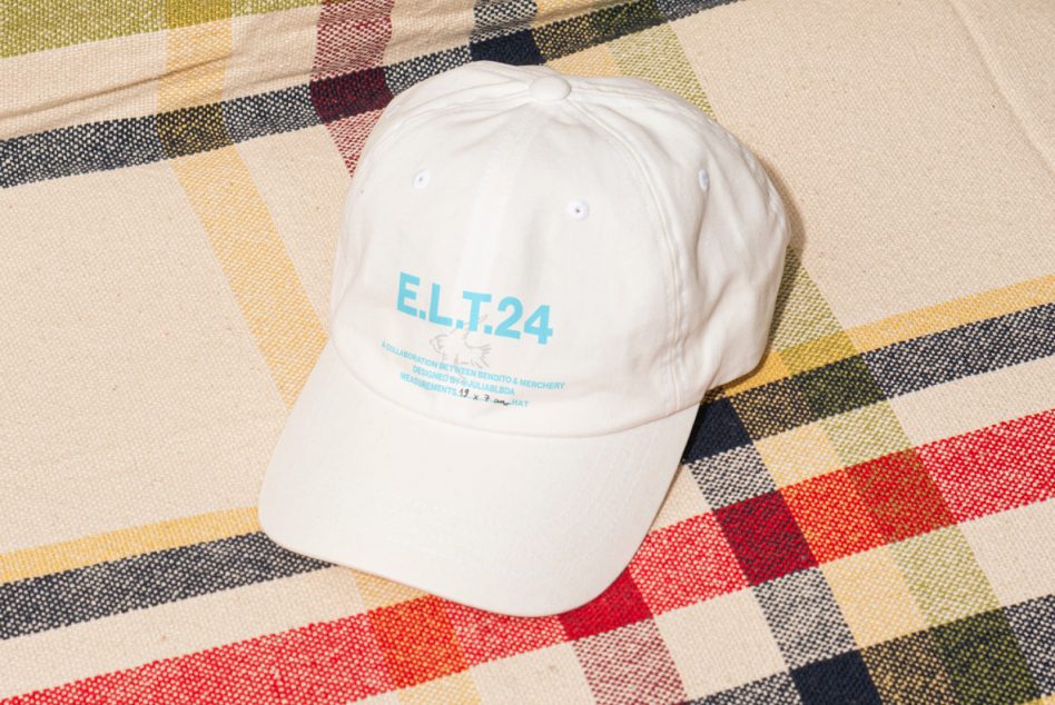 White baseball cap with blue E.L.T.24 lettering, displayed on a multicolored plaid fabric, ideal for clothing mockups, product visualization graphics