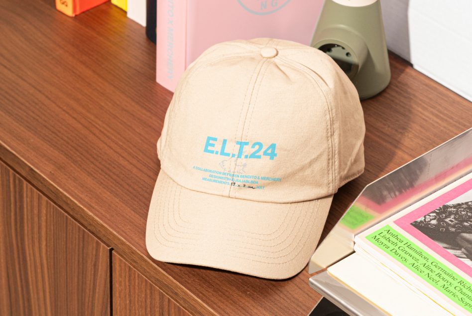 Beige cap with blue E.L.T.24 print placed on wooden desk with books and decorative objects; ideal for showcasing apparel graphic designs in digital mockups.