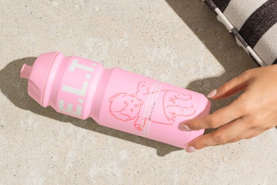 Pink water bottle with E.L.T. lettering, child cartoon graphic. Pink bike accessory held by one hand on pavement. Mockups, templates, graphic design asset.