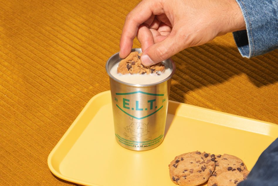 Hand dipping chocolate chip cookie into creamy drink in metallic can on yellow tray