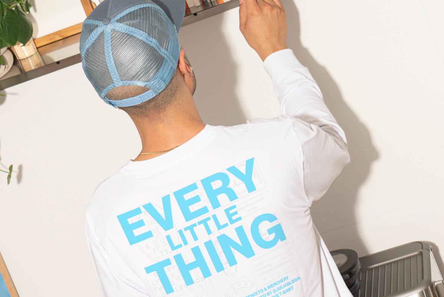 Rear view of man in white long-sleeve shirt with blue text reading Every Little Thing, wearing a blue cap, reaching up in modern interior space keywords Mockups, Templates, Design.