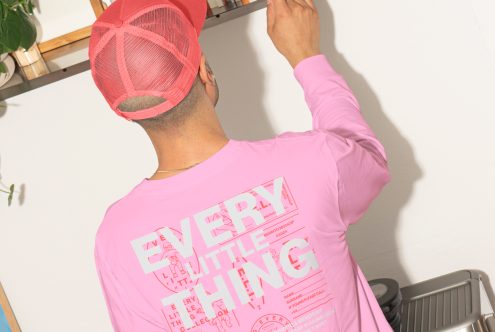 Man in pink shirt mockup with back design text reading Every Little Thing adjusting items on the shelf. Modern apparel design. Ideal for digital mockup templates.