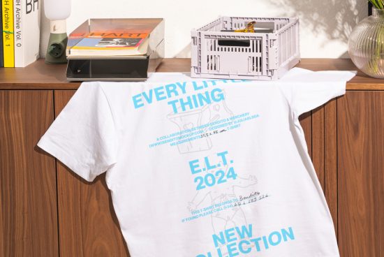 White graphic t-shirt mockup draped over a wooden cabinet with books, lamp, and storage crate on top. Perfect for showcasing t-shirt designs for designers.