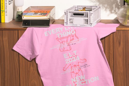 Pink T-shirt mockup featuring typography and illustrations, draped over a wooden cabinet with books and a crate. Ideal for showcasing graphic designs.