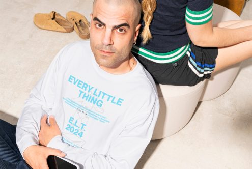 Man in white long-sleeve graphic shirt holding phone, sitting on floor next to person in shorts and sweater. Casual apparel, lifestyle, branding mockup.