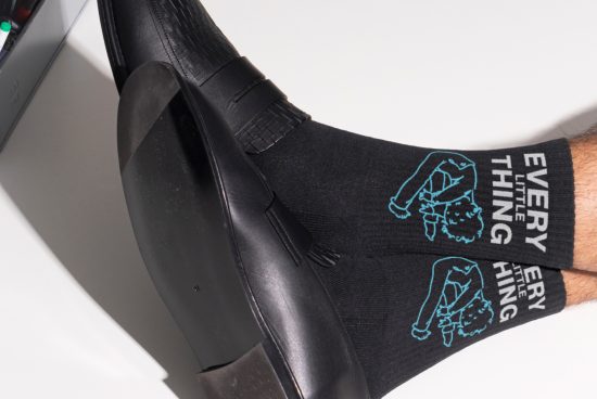 Black socks with a blue design and the text Every Little Thing worn with black shoes, ideal for mockups or graphic design templates for clothing.