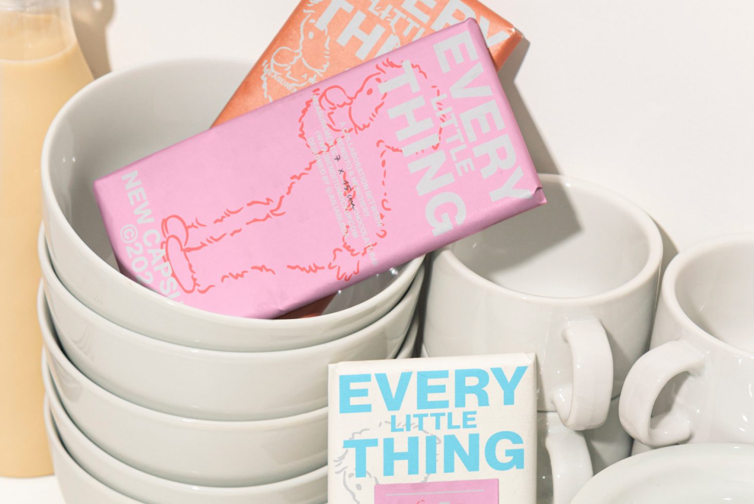 Stacked white bowls and coffee cups with pink and orange packaging of designer products labelled Every Little Thing. Great for mockups, templates, and branding.