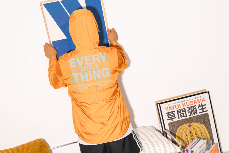 Designer in an orange jacket hanging geometric art on a wall. Books and a framed Yayoi Kusama print rest on a chair. Keywords: mockups, wall art, design, illustration.