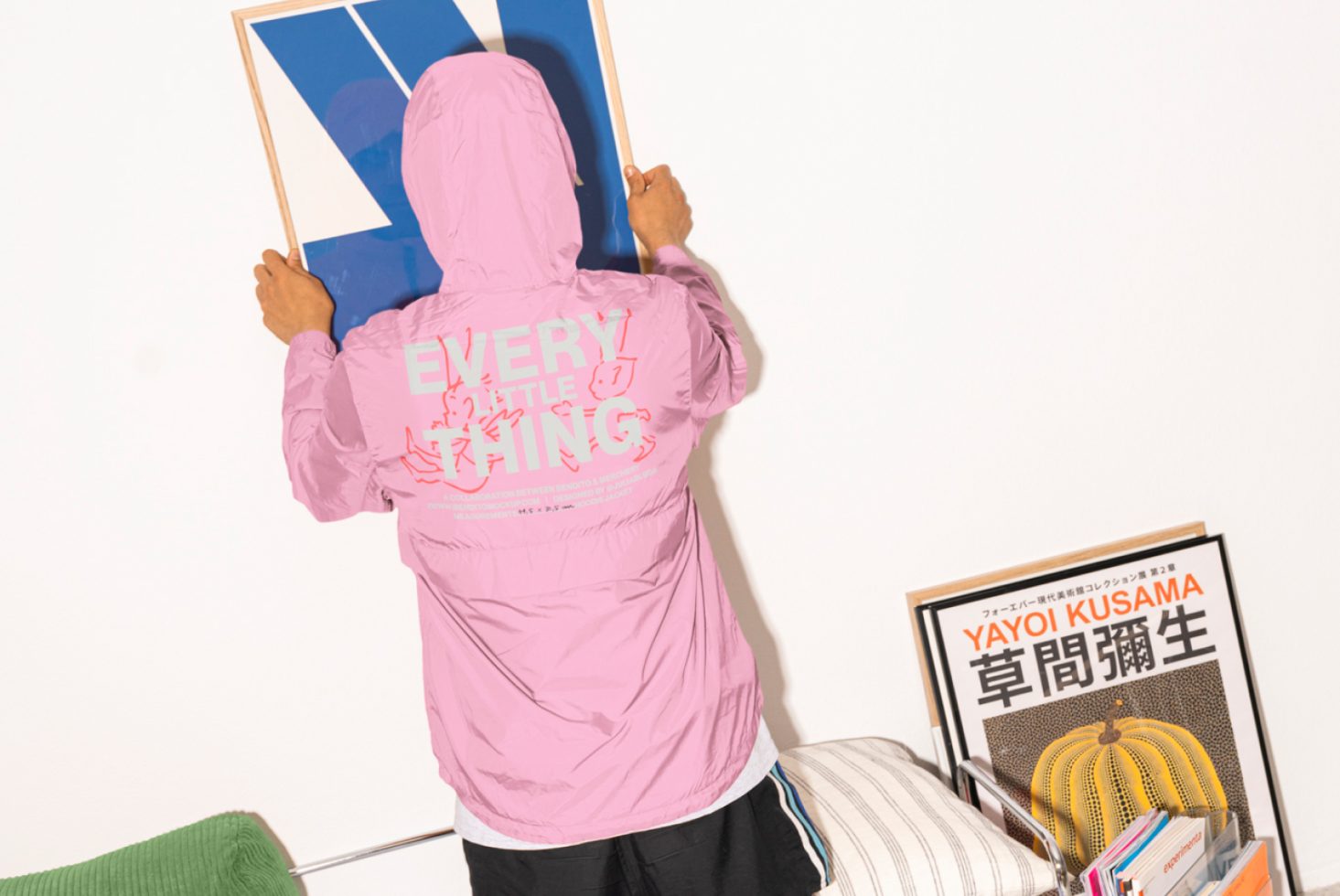 Person in pink hoodie hangs blue geometric art while facing wall. Typography print on hoodie. Yayoi Kusama book on shelf. Design inspiration for graphics.