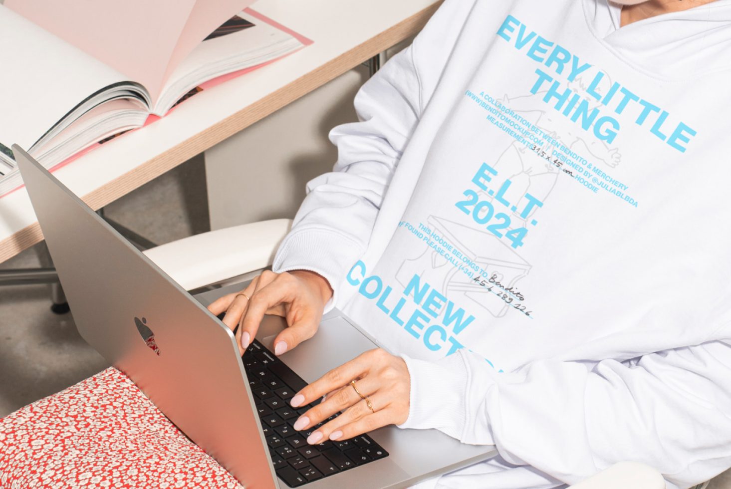 Person wearing a white hoodie working on a laptop at a desk with open books. Great mockup resource for apparel designers developing new collections.