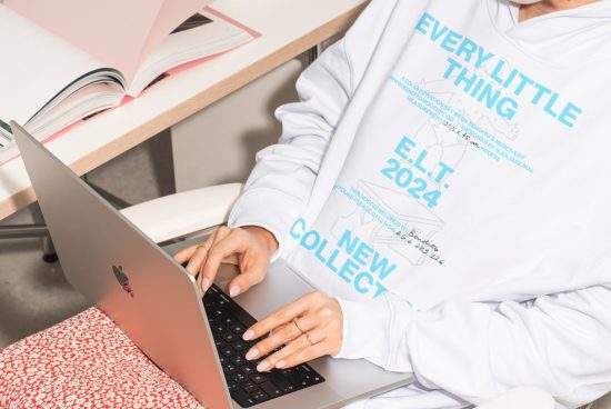 Person wearing a white hoodie working on a laptop at a desk with open books. Great mockup resource for apparel designers developing new collections.