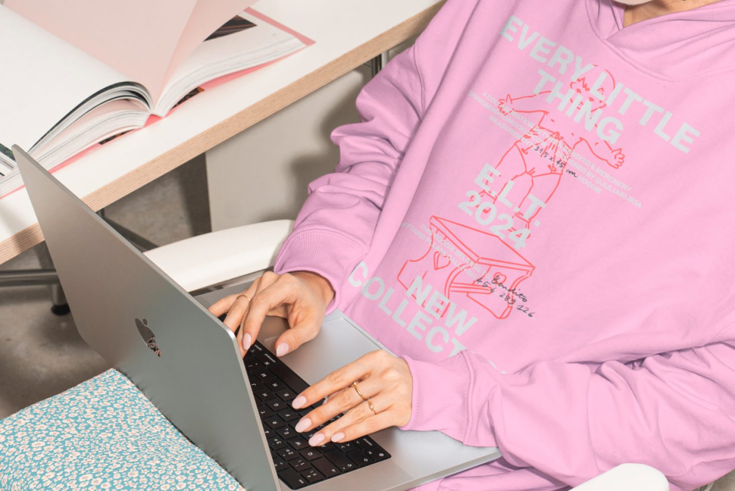 Person wearing pink hoodie typing on laptop with open books on desk. Digital mockup design, workspace inspiration, designer tools. Creative workspace setup