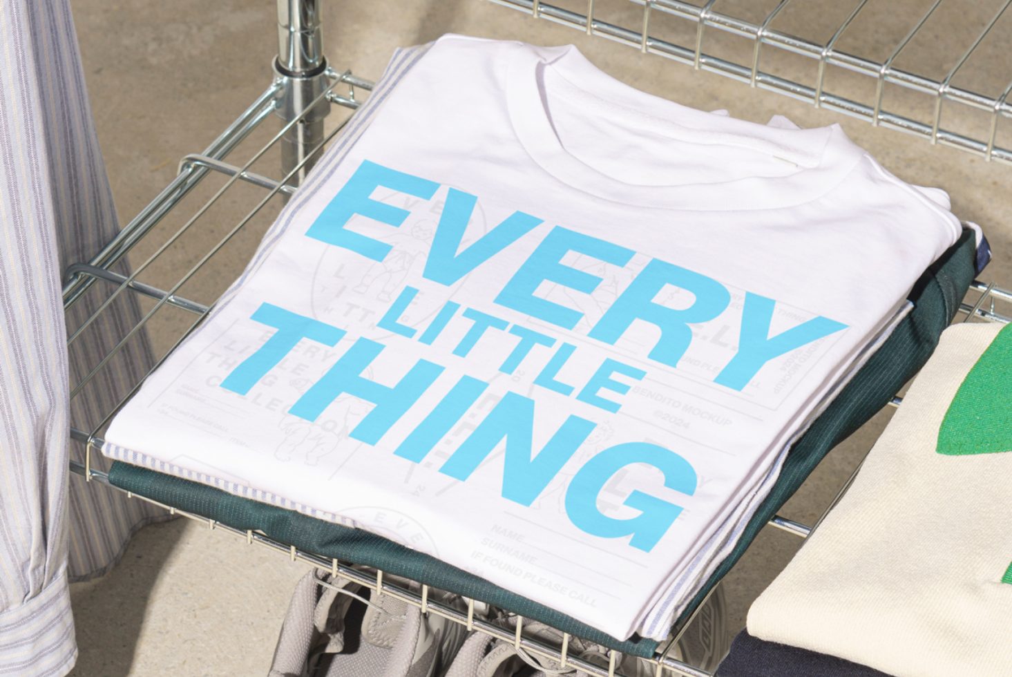 Folded white T-shirt with bold blue text EVERY LITTLE THING on it placed on a metal rack. Perfect for apparel mockups, T-shirt designs, and graphic templates for designers.