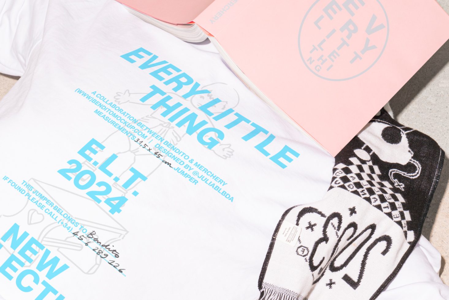 White t-shirt with blue text Every Little Thing 2024 E.L.T on it, lying next to a black and white checker scarf and a pink book. Mockup for designers.