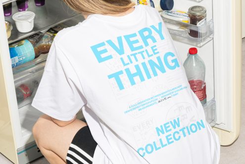 Person wearing white t-shirt with blue text Every Little Thing New Collection crouching in front of an open refrigerator. Perfect for apparel mockup templates.