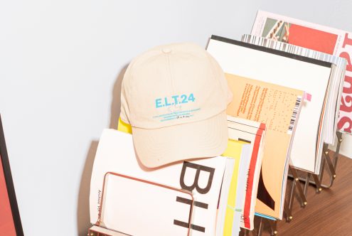 Beige cap with blue text E.L.T.24 on a wire magazine rack holding colorful magazines wood surface. Keywords mockups, templates, graphics, design, magazines.