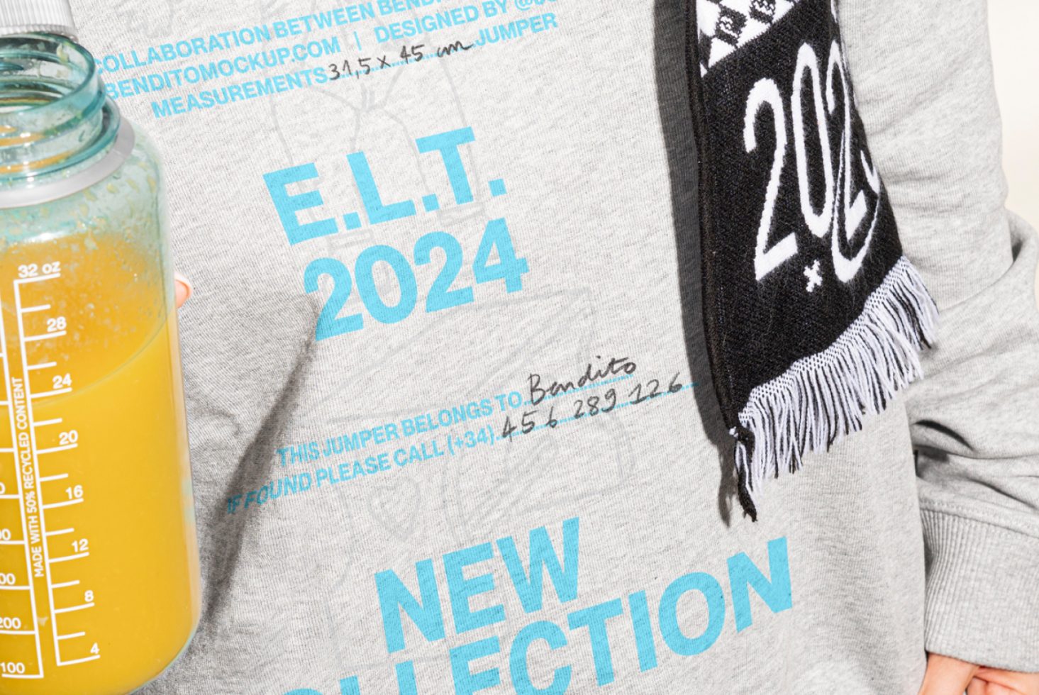 Close-up of a gray jumper featuring blue typography for a 2024 collection mockup worn with a black and white scarf and holding a bottle with orange juice.