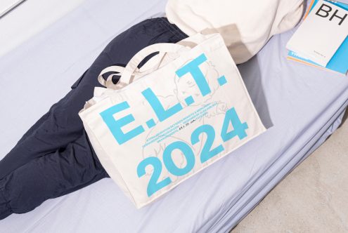 Mockup of a tote bag with bold turquoise text E.L.T. 2024 laying on a bed next to a person and books. Ideal for showcasing designs in graphics and templates.