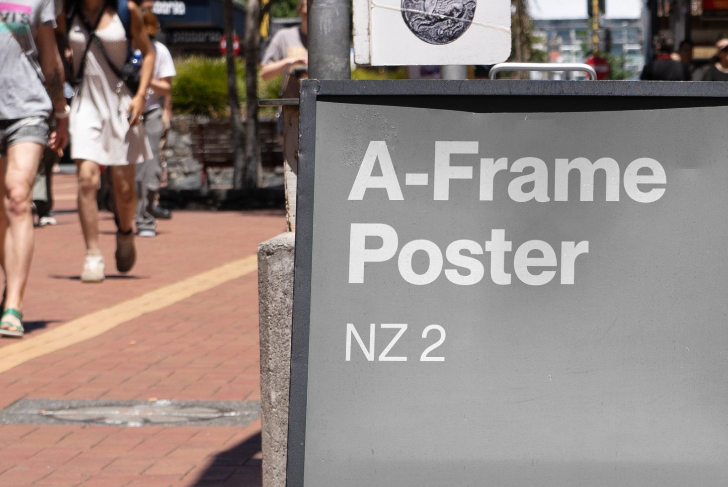 Urban street with people walking past a large A-frame poster sign reading A-Frame Poster NZ 2. Ideal for designers seeking signage mockup templates.