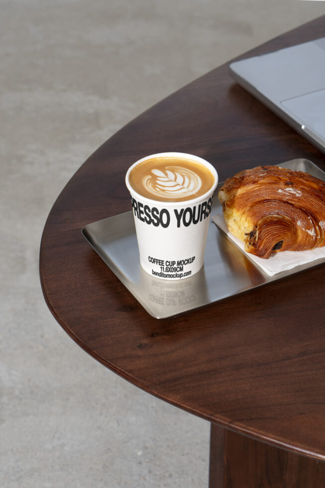 Coffee cup mockup featuring latte art design placed next to a croissant on a tray on a wooden table, ideal for showcasing branding in cafe themes.