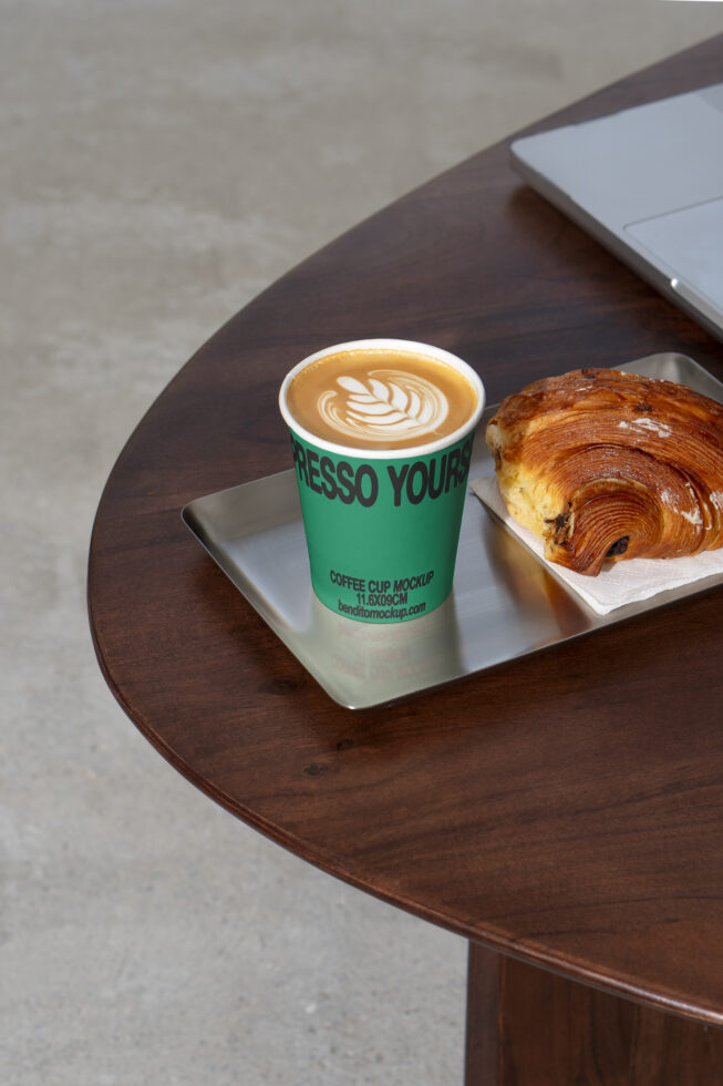 Coffee cup mockup featuring a latte with latte art and a pastry on a metal tray on a dark wooden table ideal for designers seeking realistic visuals.