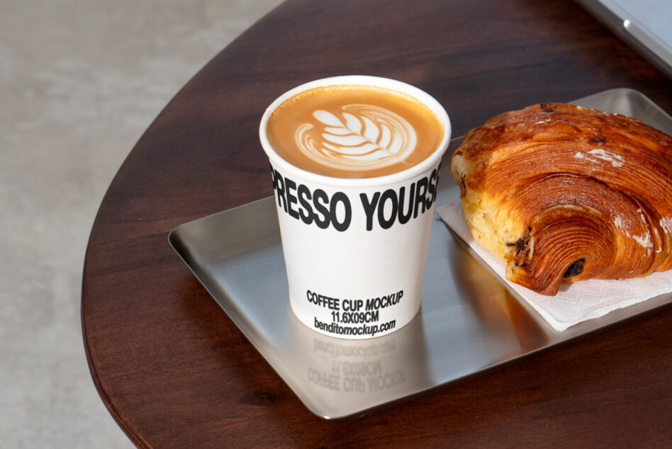 Coffee cup mockup with latte art next to a croissant on a tray. Perfect for designers seeking high-quality cafe-inspired design assets.