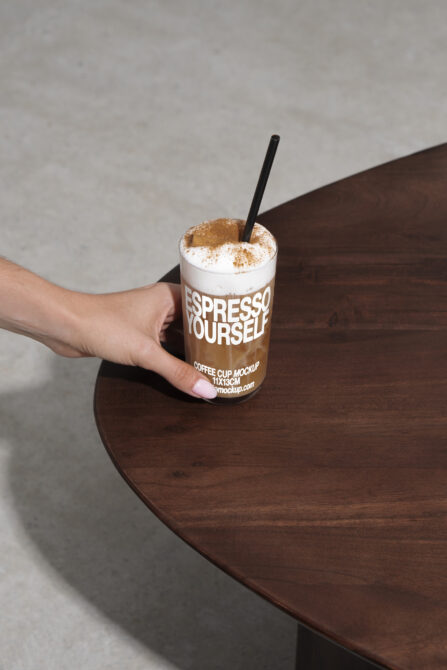 Coffee cup mockup with foam and cinnamon held by a hand over a wooden table. Perfect for designers looking for realistic beverage branding assets.