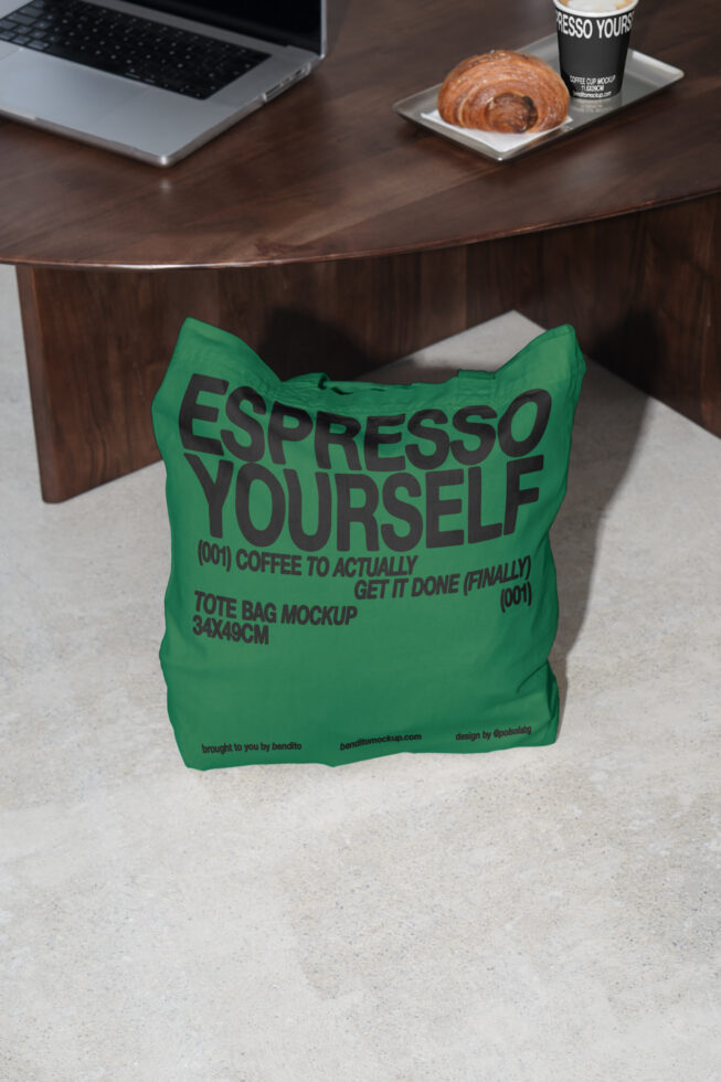 Green tote bag mockup with Espresso Yourself text. Designers product for showcasing graphics. Features laptop, coffee cup, croissant on wood table.