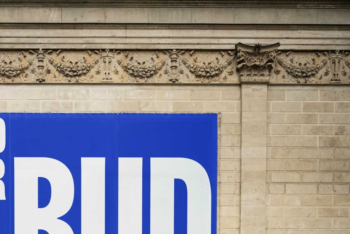 Architectural stone frieze with ornate floral details above a bold blue advertisement on a brick wall. Ideal for mockups and graphic design projects.