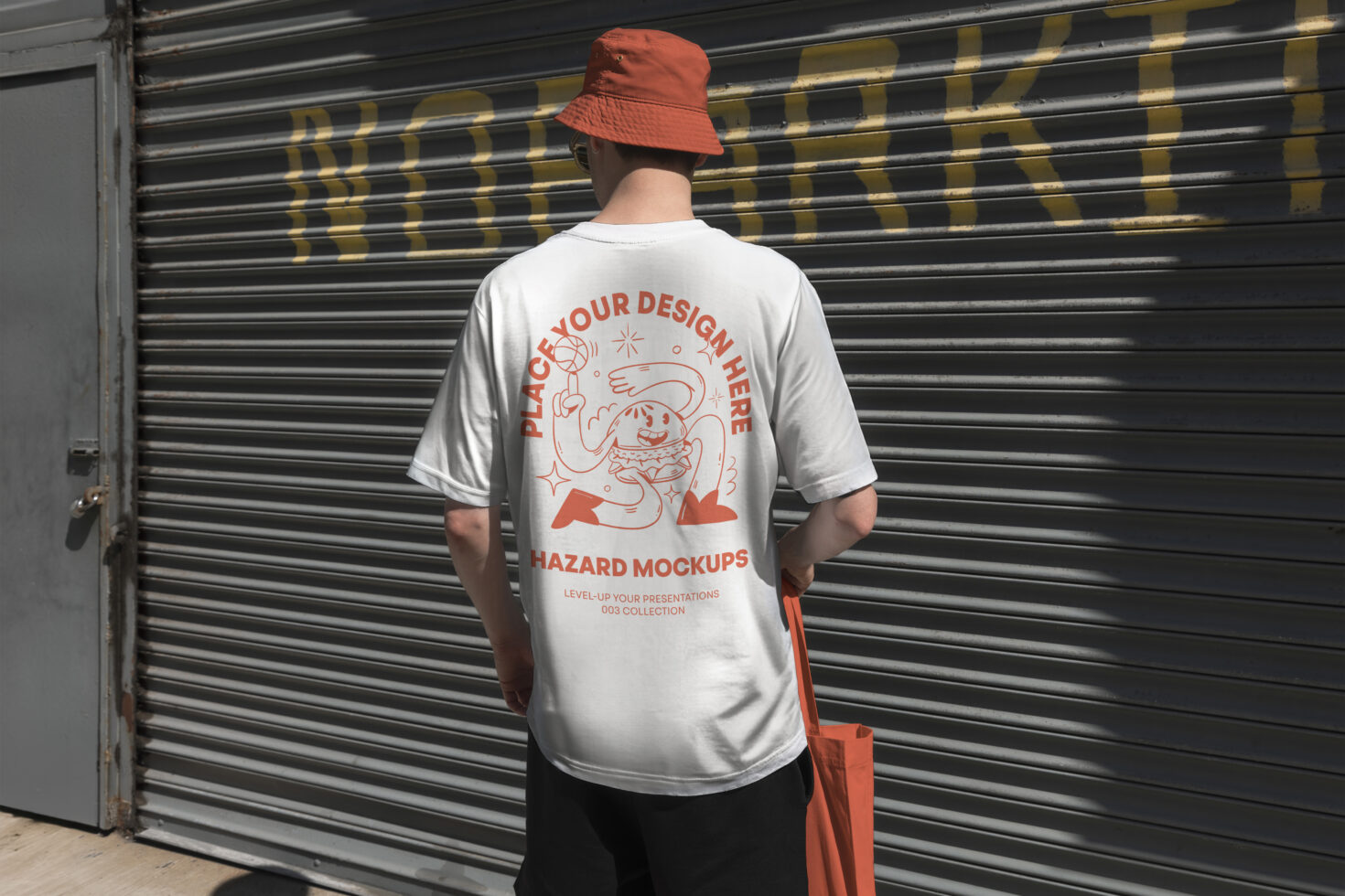 Man wearing a custom mockup t-shirt with graphic design print holding orange tote bag standing in front of garage door. Ideal for designers presentations.