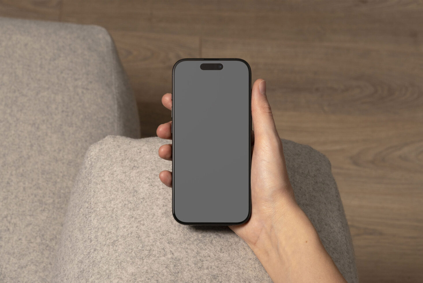 Hand holding smartphone with blank screen mockup on a gray fabric sofa. Ideal for app design, UX/UI showcase, mobile template, digital assets.