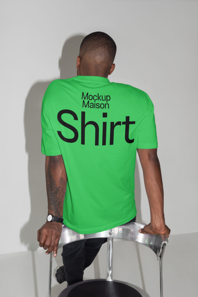 Back view of person in green mockup shirt sitting on a chair in designer studio setting. Ideal for digital assets. Keywords: mockup, shirt, template.