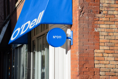 Blue storefront awning with white typography and a matching round sign displaying No 011 against a red brick wall. Ideal for branding mockups and designers.