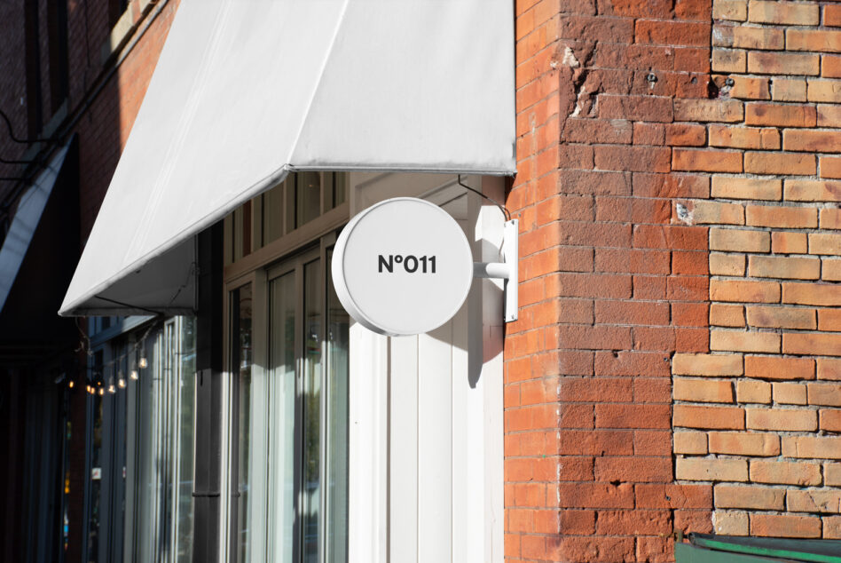 Round mockup sign on a brick wall with white storefront awning day scene. Ideal for designers. Keywords include mockup sign, storefront, branding.