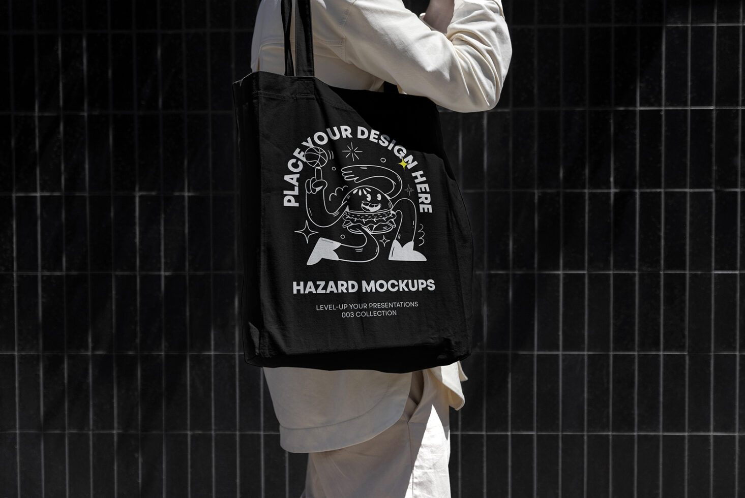 Mockup of a black tote bag with customizable design graphic and text held by a person in white against a dark tiled background. Ideal for designers.