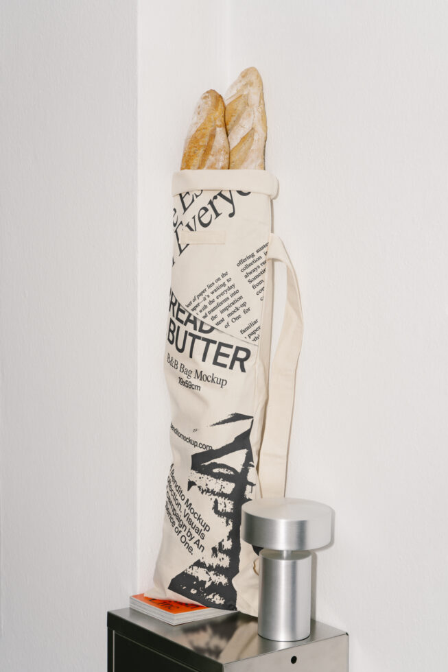 Bread bag mockup featuring artisan baguettes in a printed fabric bag placed next to a modern metal lamp on a minimalist corner shelf. Ideal for designers.