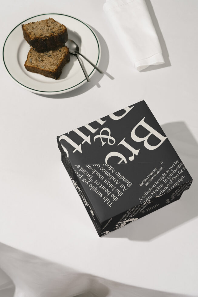 Box packaging mockup on table with rustic theme. Includes a black box with bold white text near a plate with cake slices. Ideal for branding designs.