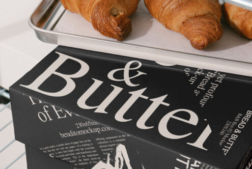 Modern packaging design mockup featuring black box with bold serif typography that reads Bread & Butter; croissants on metal tray in background.