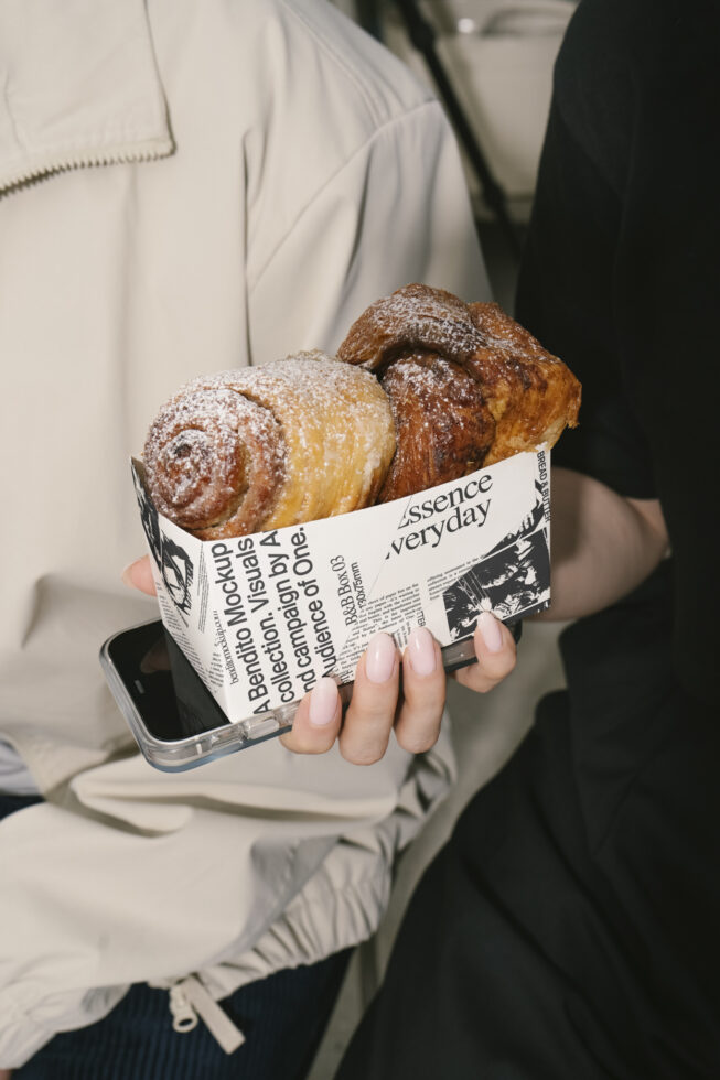 Hand holding a box of pastries wrapped in newspaper print mockup, ideal for graphic design and packaging templates, with phone visible underneath.