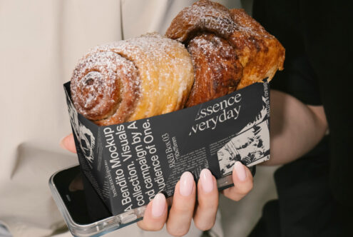 A hand holding a black paper mockup with pastries atop a phone. Perfect for designers seeking modern packaging design. Keywords: Mockup, Packaging Design.