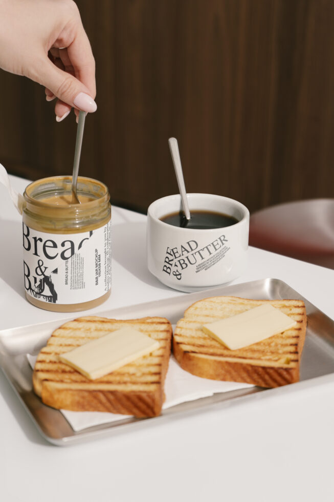 Coffee and toast branding mockup on a table with a hand holding a spoon in a jar next to a cup displaying bread and butter design, suited for designers.