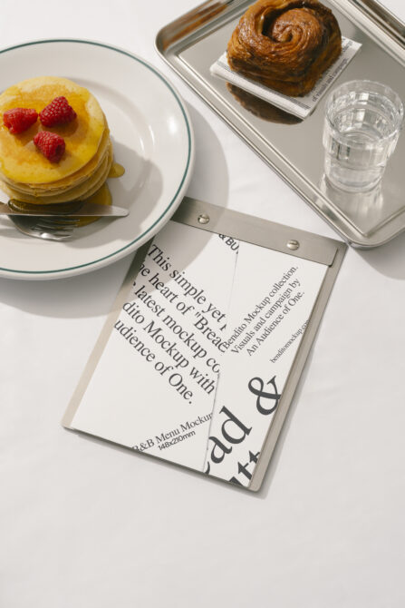 Menu mockup with elegant typography alongside pancakes with raspberries and a pastry on a tray. Perfect for restaurant branding or design presentations.