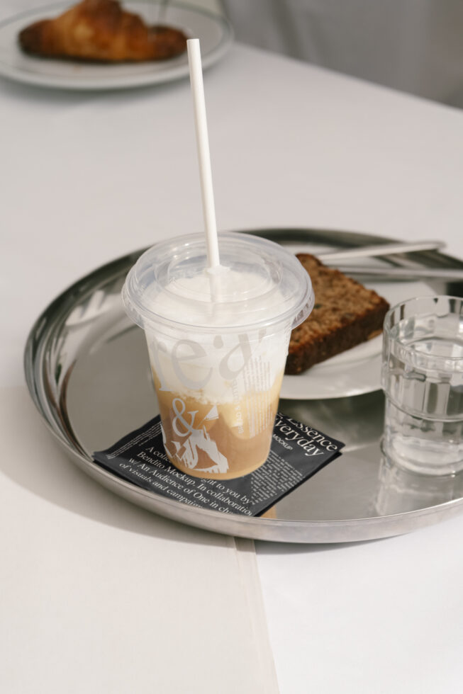 Cafe branding mockup featuring an iced coffee in a branded plastic cup on a silver tray with a croissant and water. Ideal for designers seeking stylish graphics.