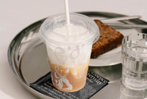 Mockup of a transparent plastic cup with straw featuring smoothie on a metal tray with a slice of bread and glass of water on designer tabletop setting.